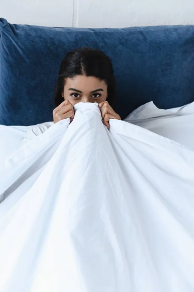 Hermosa chica de raza mixta mirando desde la manta en la mañana en el dormitorio y mirando a la cámara - foto de stock