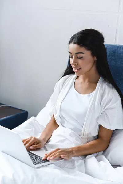 Sorrindo atraente menina de raça mista em roupão branco usando laptop de manhã na cama — Fotografia de Stock