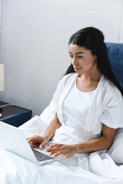 Attractive mixed race girl in white robe using laptop in morning in bed — Stock Photo