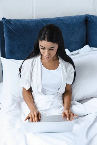 Vista de ángulo alto de la hermosa chica de raza mixta en bata blanca utilizando el ordenador portátil en la mañana en la cama - foto de stock