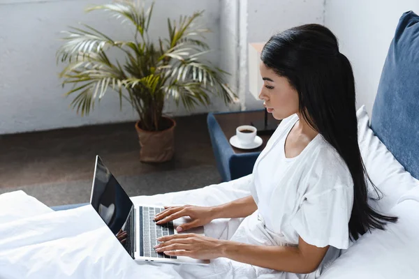 High angle view of beautiful mixed race girl in white robe using laptop in morning in bedroom — Stock Photo