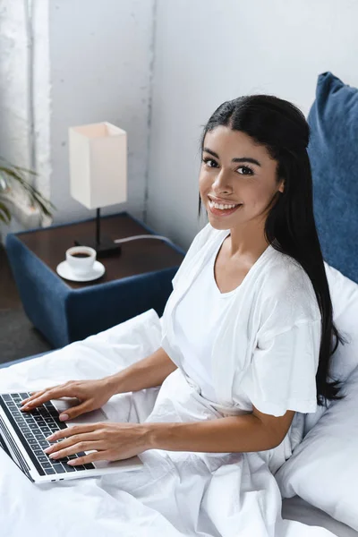 Vista ad alto angolo di sorridere bella ragazza razza mista in accappatoio bianco utilizzando il computer portatile al mattino in camera da letto e guardando la fotocamera — Foto stock