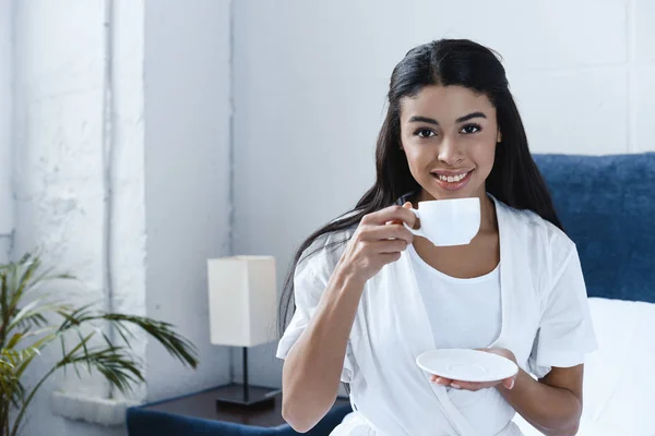 Lächelnd schönes Mischlingsmädchen in weißem Gewand, das Kaffee trinkt und morgens im Schlafzimmer in die Kamera schaut — Stockfoto