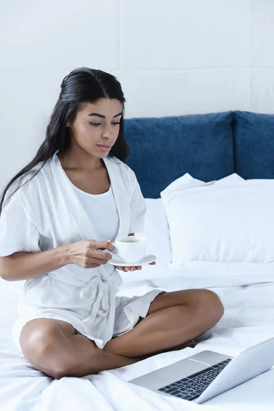 Hermosa chica de raza mixta en túnica blanca sosteniendo taza de café y mirando a la computadora portátil en la mañana en el dormitorio - foto de stock