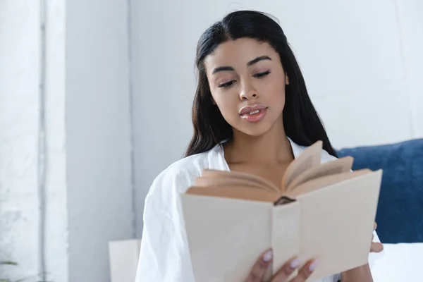 Atraente menina de raça mista em robe branco leitura livro na cama de manhã — Fotografia de Stock