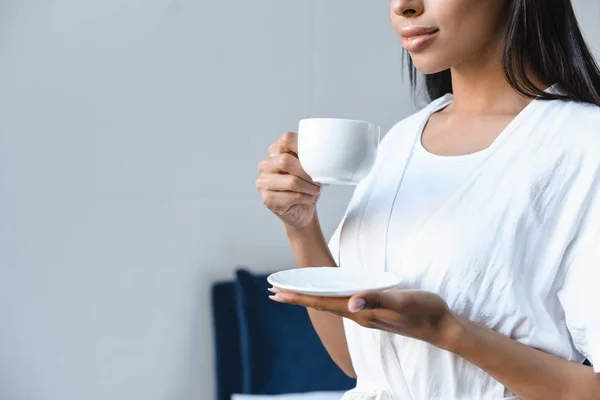 Imagem cortada de menina de raça mista em roupão branco segurando xícara de café de manhã no quarto — Fotografia de Stock