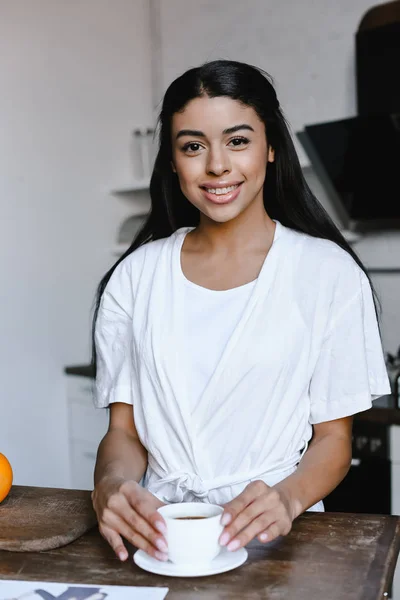 Sonriente hermosa chica de raza mixta en túnica blanca sosteniendo taza de café y mirando a la cámara en la mañana en la cocina - foto de stock