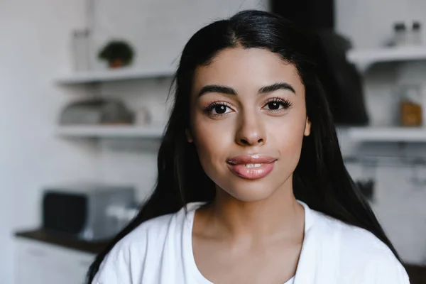 Retrato de hermosa chica de raza mixta en bata blanca mirando a la cámara en la mañana en la cocina - foto de stock