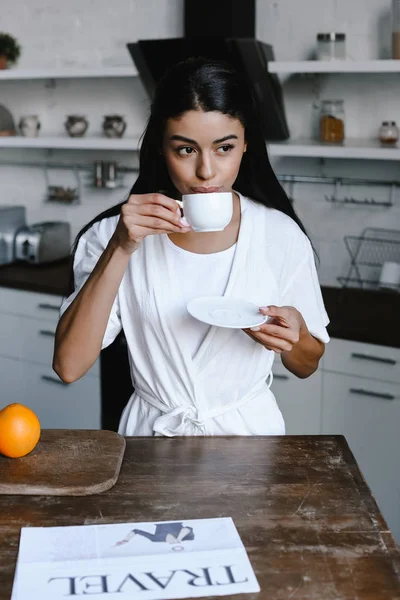 Schönes Mischlingsmädchen in weißem Gewand, das morgens in der Küche Kaffee trinkt — Stockfoto