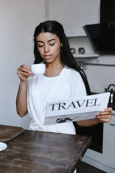 Bela menina de raça mista em roupão branco segurando xícara de café e lendo jornal de viagem de manhã na cozinha — Fotografia de Stock