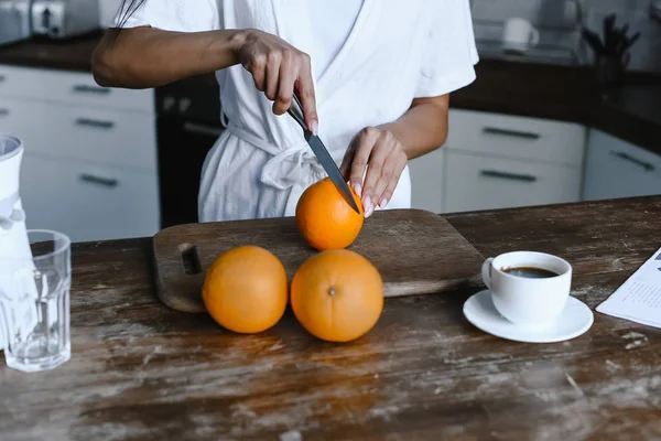 Abgeschnittenes Bild von Mischlingsmädchen in weißer Robe, die morgens in der Küche Orangen schneidet — Stockfoto