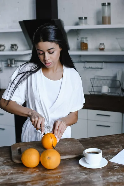 Schönes Mischlingsmädchen in weißer Robe bereitet Orangensaft am Morgen zu und schneidet Orangen in der Küche — Stockfoto