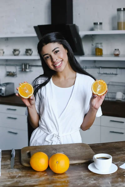 Sourire belle mixte fille en robe blanche tenant coupé orange et regardant la caméra le matin dans la cuisine — Photo de stock