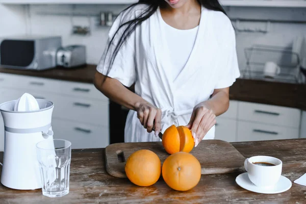 Imagem cortada de menina de raça mista em roupão branco cortando laranjas maduras de manhã na cozinha — Fotografia de Stock