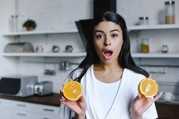 Shocked mixed race girl in white robe holding cut orange and looking at camera in morning in kitchen — Stock Photo