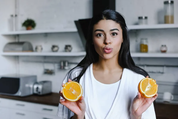 Beautiful mixed race girl in white robe holding cut orange, grimacing and looking at camera in morning in kitchen — Stock Photo