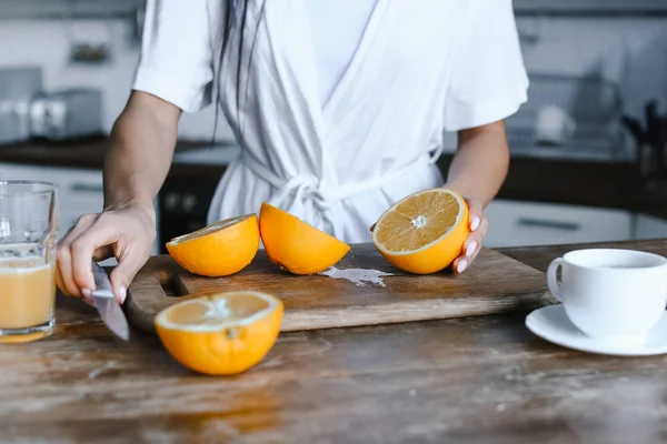Обрезанный образ девушки смешанной расы в белом халате, готовящей апельсиновый сок утром и режущей апельсины на кухне — стоковое фото