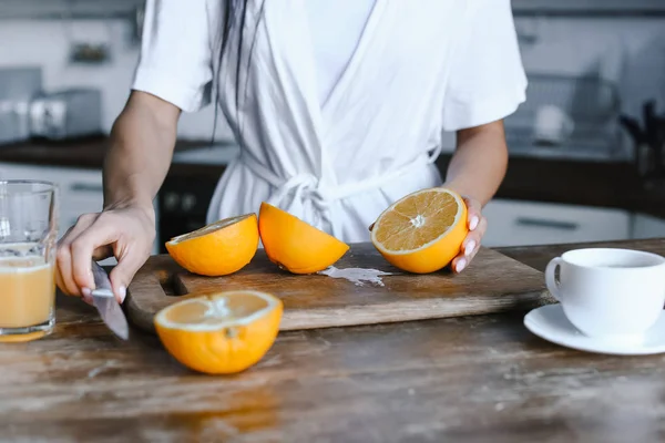 Imagen recortada de chica de raza mixta en bata blanca preparando jugo de naranja por la mañana y el corte de naranjas en la cocina — Stock Photo