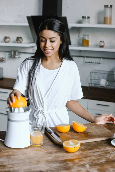 Sorridente bella ragazza razza mista in accappatoio bianco preparare succo d'arancia al mattino in cucina — Foto stock