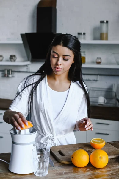 Hermosa chica de raza mixta en bata blanca preparando jugo de naranja en la mañana en la cocina - foto de stock