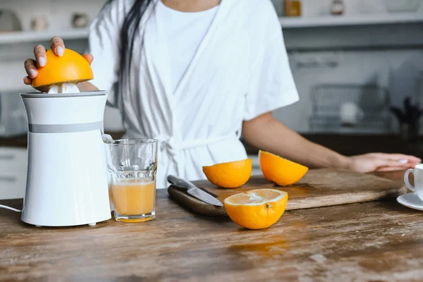 Cropped image of mixed race girl in white robe preparing orange juice in morning in kitchen — Stock Photo