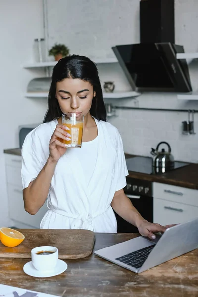 Beautiful mixed race girl in white robe drinking juice in morning in kitchen — Stock Photo