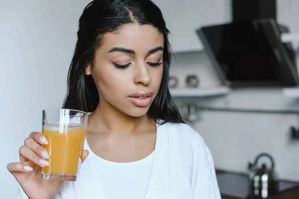 Bela menina de raça mista em roupão branco segurando vidro de suco de laranja fresco de manhã na cozinha — Fotografia de Stock