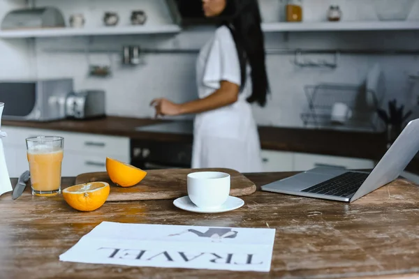 Mixed race girl in white robe in morning in kitchen, cup of coffee and travel newspaper on tabletop — Stock Photo