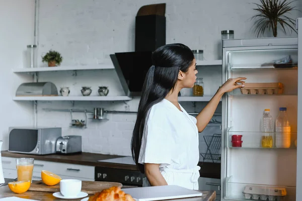 Hermosa chica de raza mixta en bata blanca por la mañana tomando huevos de la nevera en la cocina - foto de stock