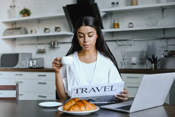Hermosa chica de raza mixta en bata blanca sosteniendo taza de café y leyendo periódico de viaje en la mañana en la cocina - foto de stock
