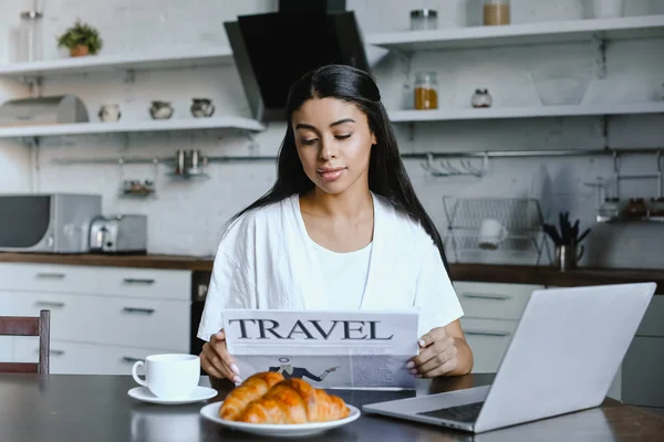 Hermosa chica de raza mixta en bata blanca leyendo periódico de viaje en la mañana en la cocina - foto de stock