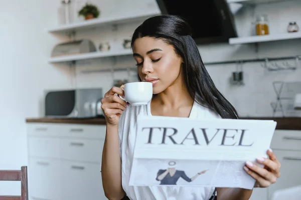 Beautiful mixed race girl in white robe drinking coffee and holding travel newspaper in morning in kitchen — Stock Photo