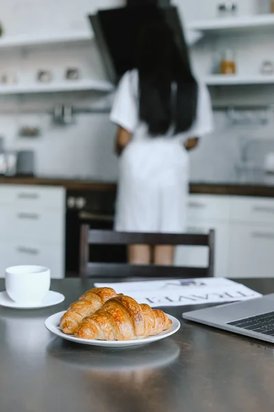 Vue arrière de la jeune fille métisse en robe blanche debout près de la cuisinière le matin dans la cuisine, croissants au premier plan — Photo de stock