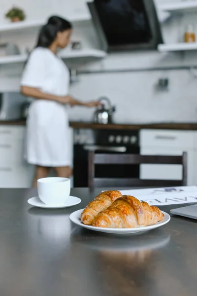 Mischlingsmädchen im weißen Gewand stellt morgens in der Küche Wasserkocher auf den Herd, Croissants im Vordergrund — Stockfoto