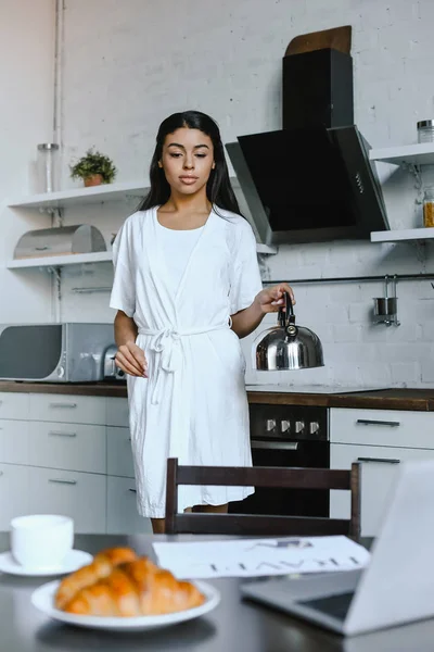 Beautiful mixed race girl in white robe holding kettle in morning in kitchen — Stock Photo