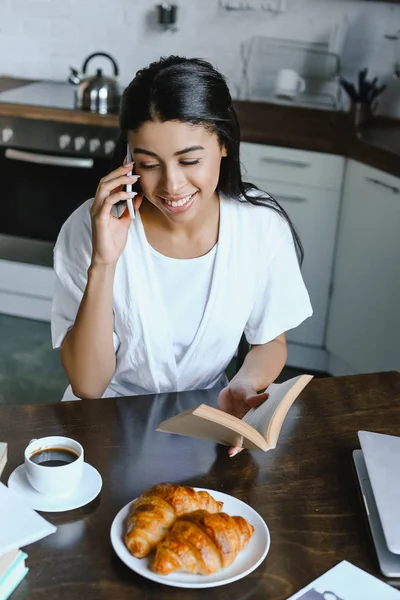 High angle view of smiling beautiful mixed race girl in white robe talking by smartphone in morning in kitchen and holding book — Stock Photo