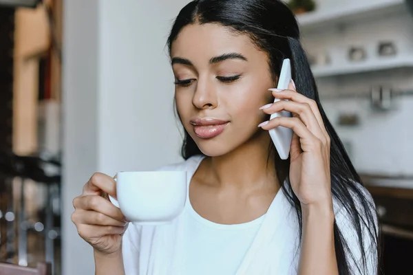 Portrait of beautiful mixed race girl in white robe talking by smartphone in morning in kitchen and drinking coffee — Stock Photo