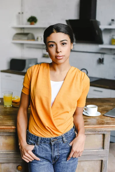 Hermosa chica de raza mixta en camisa naranja de pie en la cocina y mirando a la cámara - foto de stock