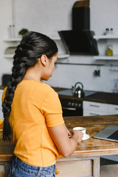 Vista trasera de chica de raza mixta en camisa naranja tomando taza de café en la cocina - foto de stock