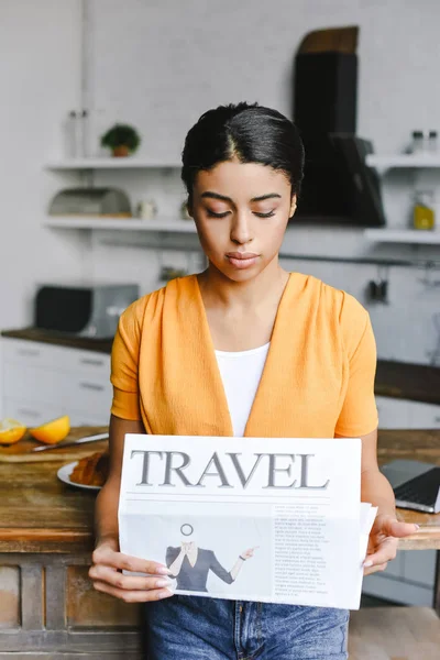 Hermosa chica de raza mixta en camisa naranja leyendo periódico de viaje en la cocina - foto de stock