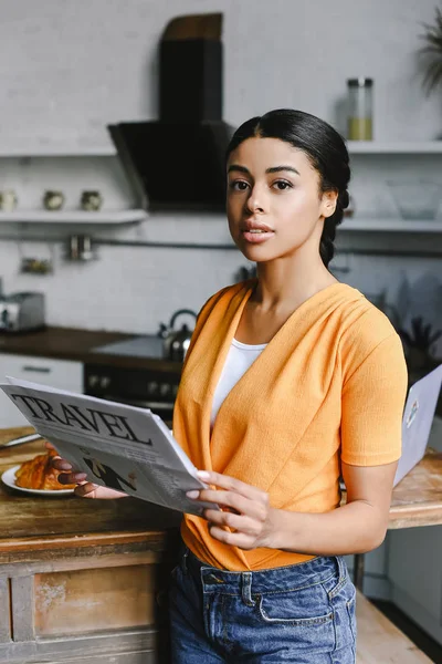 Hermosa chica de raza mixta en camisa naranja con periódico de viaje en la cocina y mirando a la cámara - foto de stock