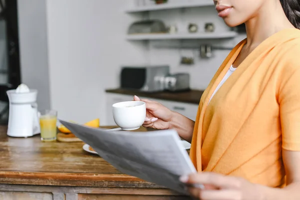 Immagine ritagliata di ragazza razza mista in camicia arancione in possesso di una tazza di caffè e lettura di giornale in cucina — Foto stock