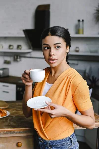 Menina de raça mista atraente na camisa laranja beber café de manhã na cozinha — Fotografia de Stock