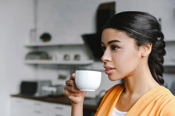 Portrait of beautiful mixed race girl in orange shirt drinking coffee in morning in kitchen — Stock Photo
