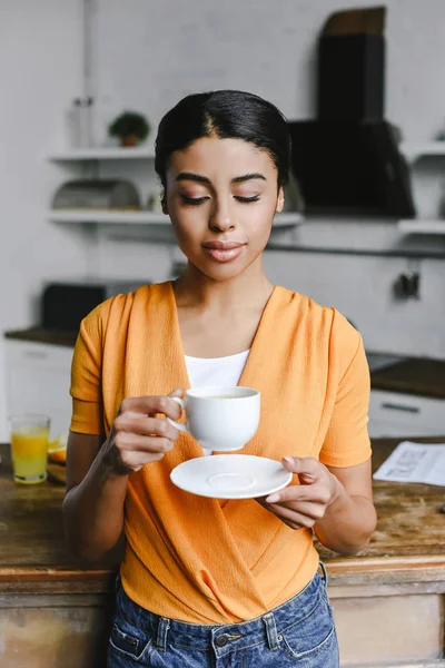 Attractive mixed race girl in orange shirt holding cup of coffee and plate in morning in kitchen — Stock Photo