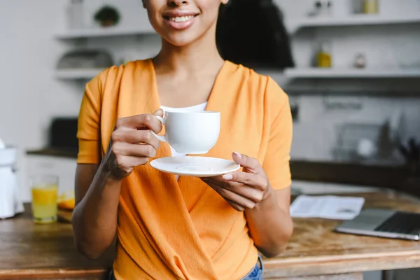 Imagen recortada de chica de raza mixta sonriente en camisa naranja sosteniendo taza de café y plato por la mañana en la cocina - foto de stock