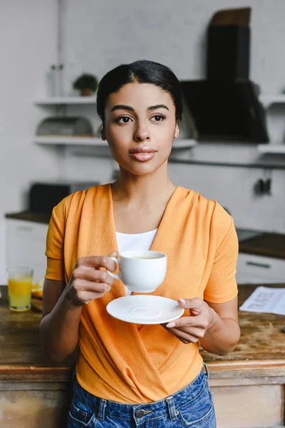 Beautiful mixed race girl in orange shirt holding cup of coffee in morning in kitchen — Stock Photo