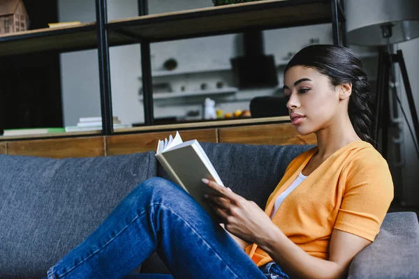 Vista lateral da bela menina de raça mista em livro de leitura camisa laranja na sala de estar — Fotografia de Stock