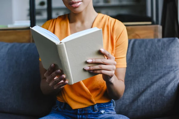 Imagen recortada de chica de raza mixta en camisa naranja libro de lectura en la sala de estar - foto de stock