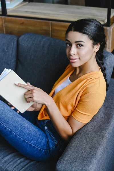 Vista de ángulo alto de hermosa chica de raza mixta en camisa naranja sosteniendo libro y mirando a la cámara en la sala de estar - foto de stock